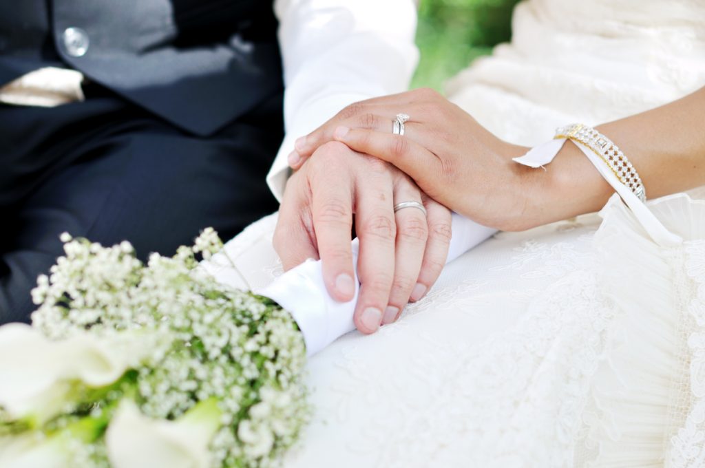 Brudepar som holder hender med giftering på hver sin finger. Etikette regler for bryllup finner du her.