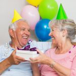 Et eldre ektepar som gir hverandre gaver med partyhatter på hodet, smilende og glade. finn en gave til 60 åring du er glad i finner du her!
