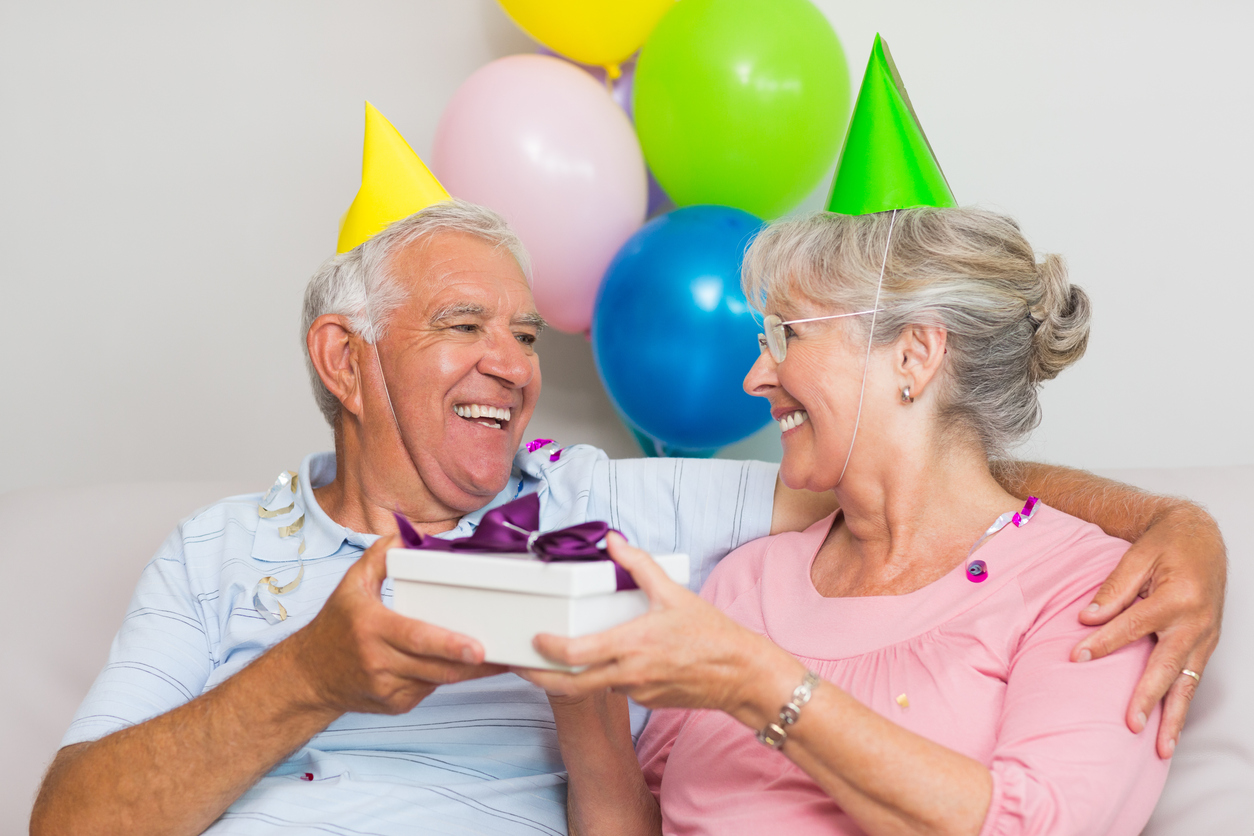 Et eldre ektepar som gir hverandre gaver med partyhatter på hodet, smilende og glade. finn en gave til 60 åring du er glad i finner du her!