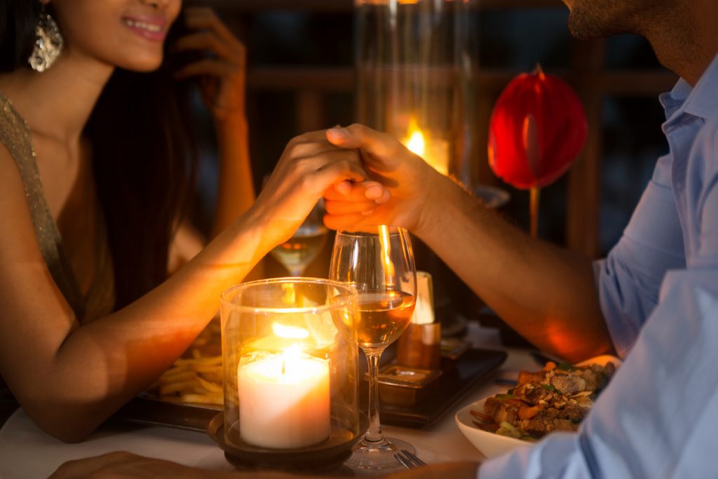 Romantisk par som holder hender under en middag med stearinlys, godt drikke i glassene og god mat. En perfekt gave til henne 2017.